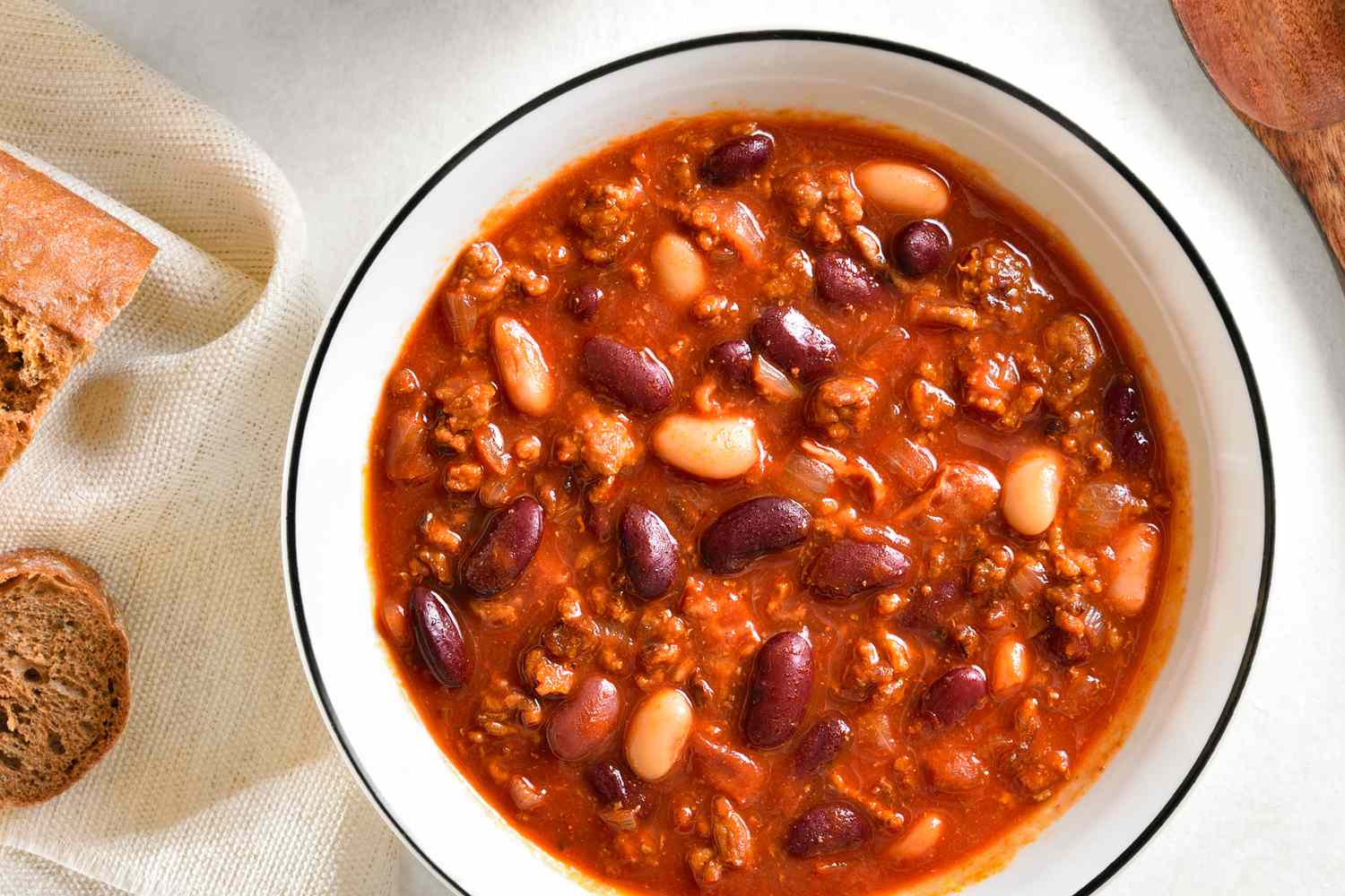 Bowl of chili with kidney and white beans on a table with bread and a spoon