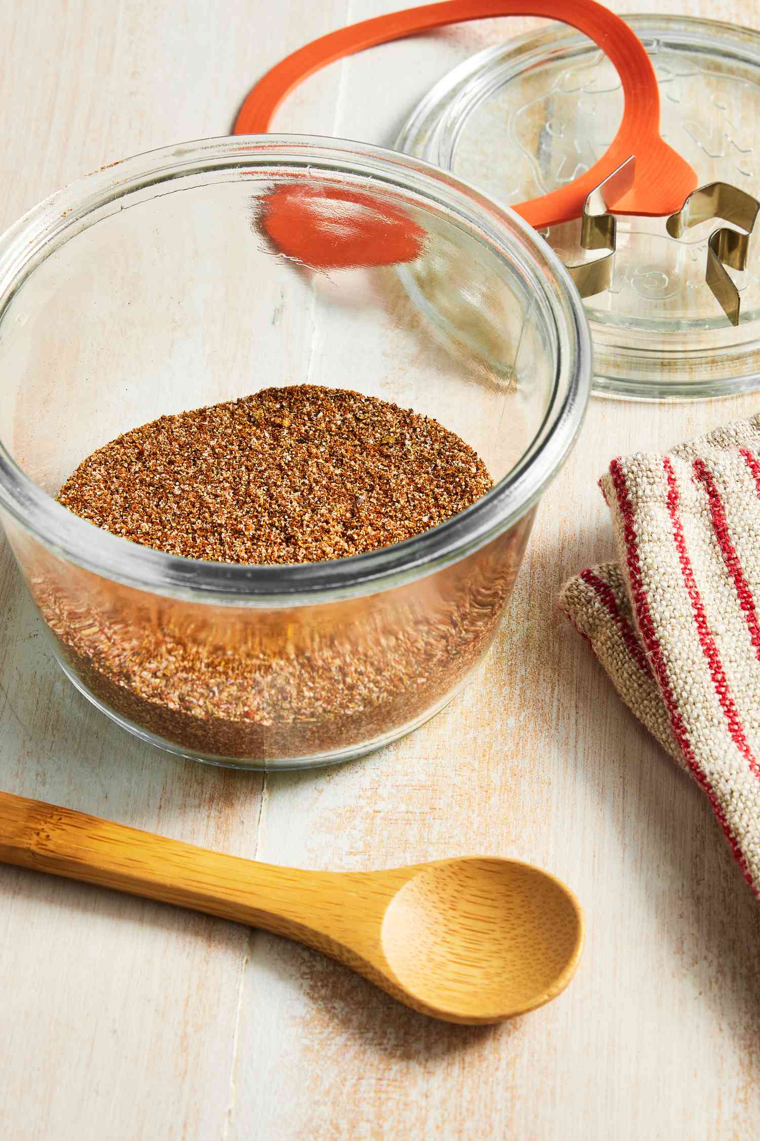 Fajita Seasoning in a Glass Jar, and in the Surroundings, a Wooden Spoon, a Red and White Stripped Kitchen Towel, and a Glass Lid for the Jar 