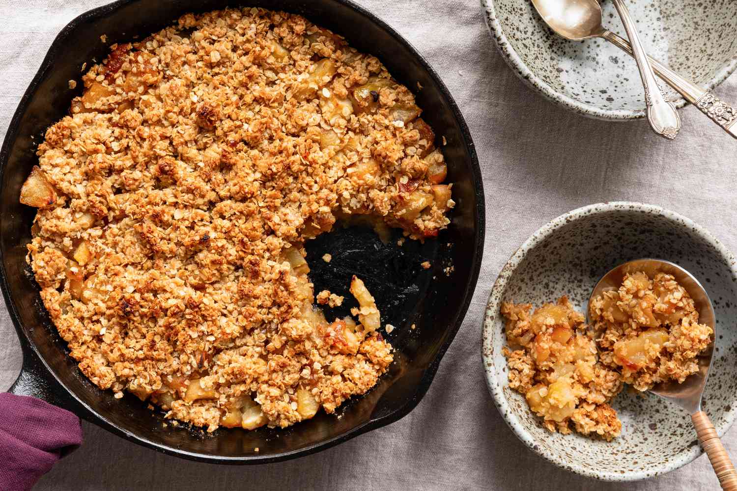 3-ingredient skillet apple crisp in a cast iron skillet and a serving in a bowl with a spoon, both next to a bowl with utensils and a purple kitchen towel