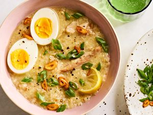 Overhead view of a pink bowl of Arroz Caldo with Chicken recipe next to a green glass and plate of lemon slices and scallions 