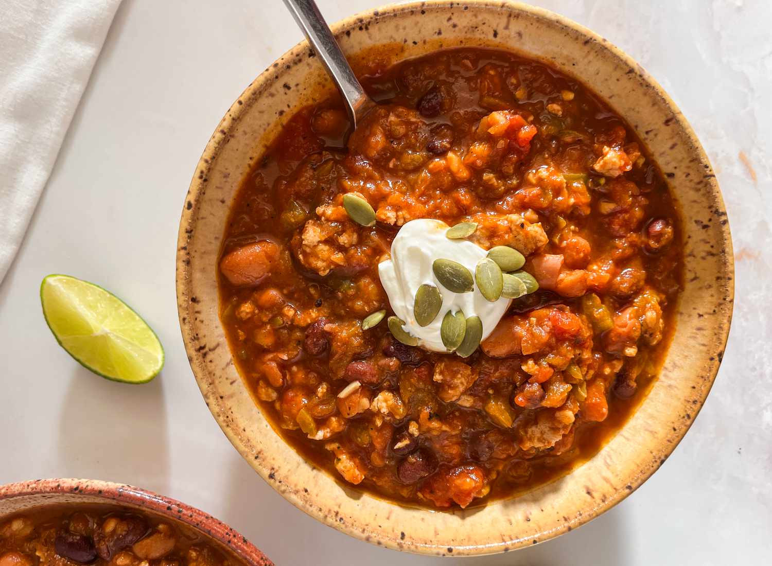 Bowl of easy pumpkin chili topped with a dollop of sour cream and pumpkin seeds at a table setting with another bowl of chili, a lime wedge on the counter, and white table napkins