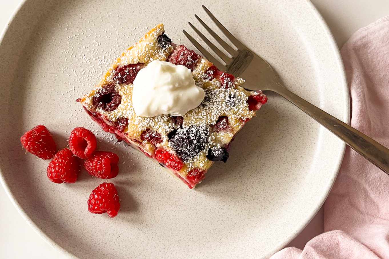 A serving of berry clafoutis topped with whipped cream on a plate with a fork, surrounded by a few raspberries
