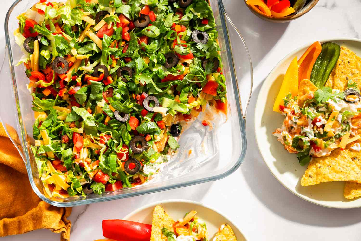 A dish of layered nacho dip with accompanying plates of chips and vegetables