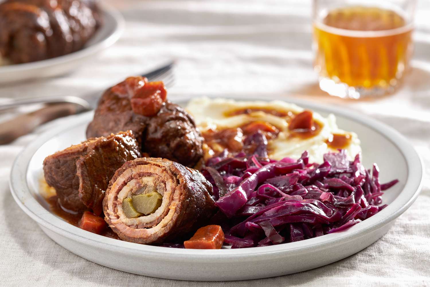 German rouladen with cabbage and mashed potatoes and gravy on a plate and in the background, a table setting with another serving on a plate and a glass of beer