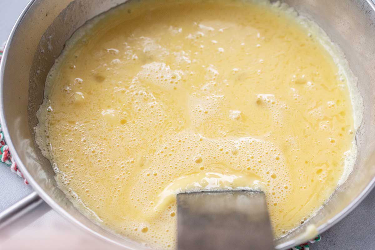 A skillet with whisked eggs covers the bottom of the pan. A spatula sits on the side of the pan.