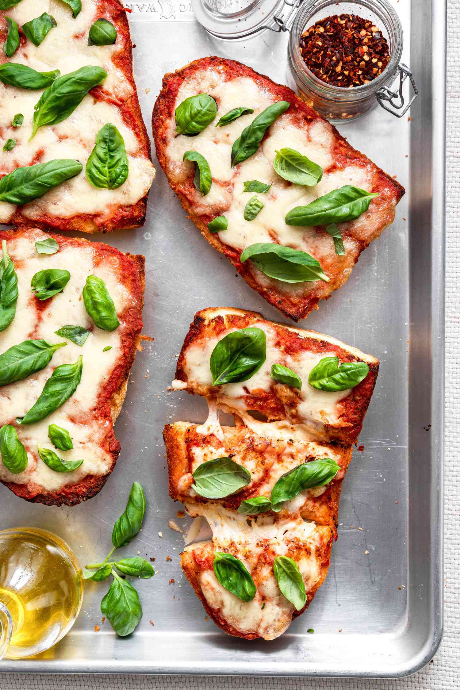 A Sliced French Bread Pizza Topped With Fresh Basil Surrounded by More Pizza (Unsliced), an Olive Oil Dispenser, and a Jar of Crushed Red Peppers on a Baking Tray