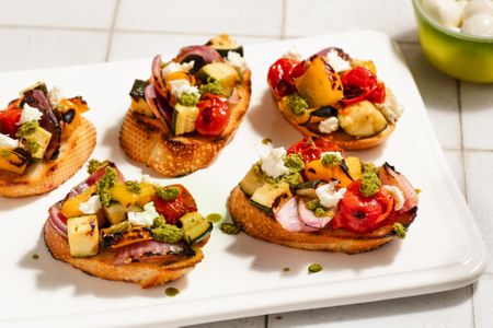 Toasts topped with grilled summer vegetables, goat cheese, and pesto on a white serving platter