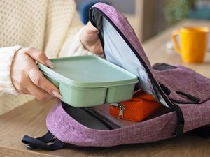 Woman wearing a white sweater putting a bento-style lunch box into a small purple backpack on a wooden tabletop