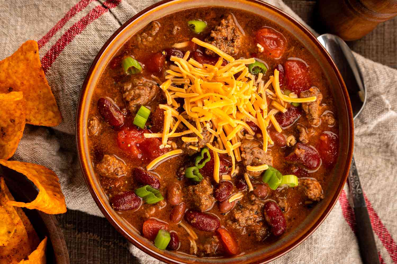 Overhead view of a brown serving bowl of chili topped with cheese and green onions sitting on a towel next to a spoon and some corn chips