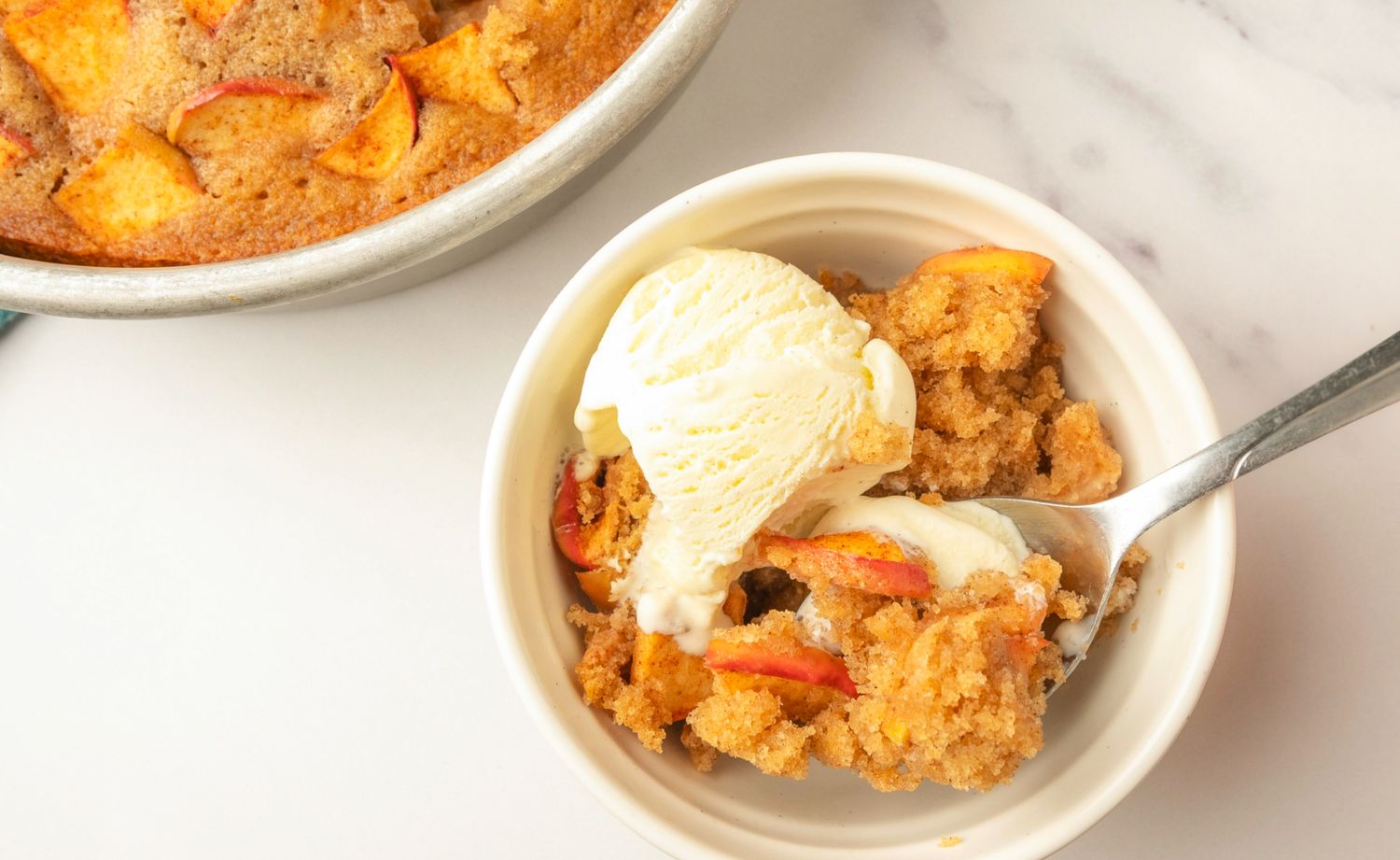 Bowl of apple spoon cake with a scoop of ice cream and a spoon pan of cake in the background