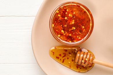 Jar of chili honey with red pepper flakes next to a honey dipper coated with the spiced honey