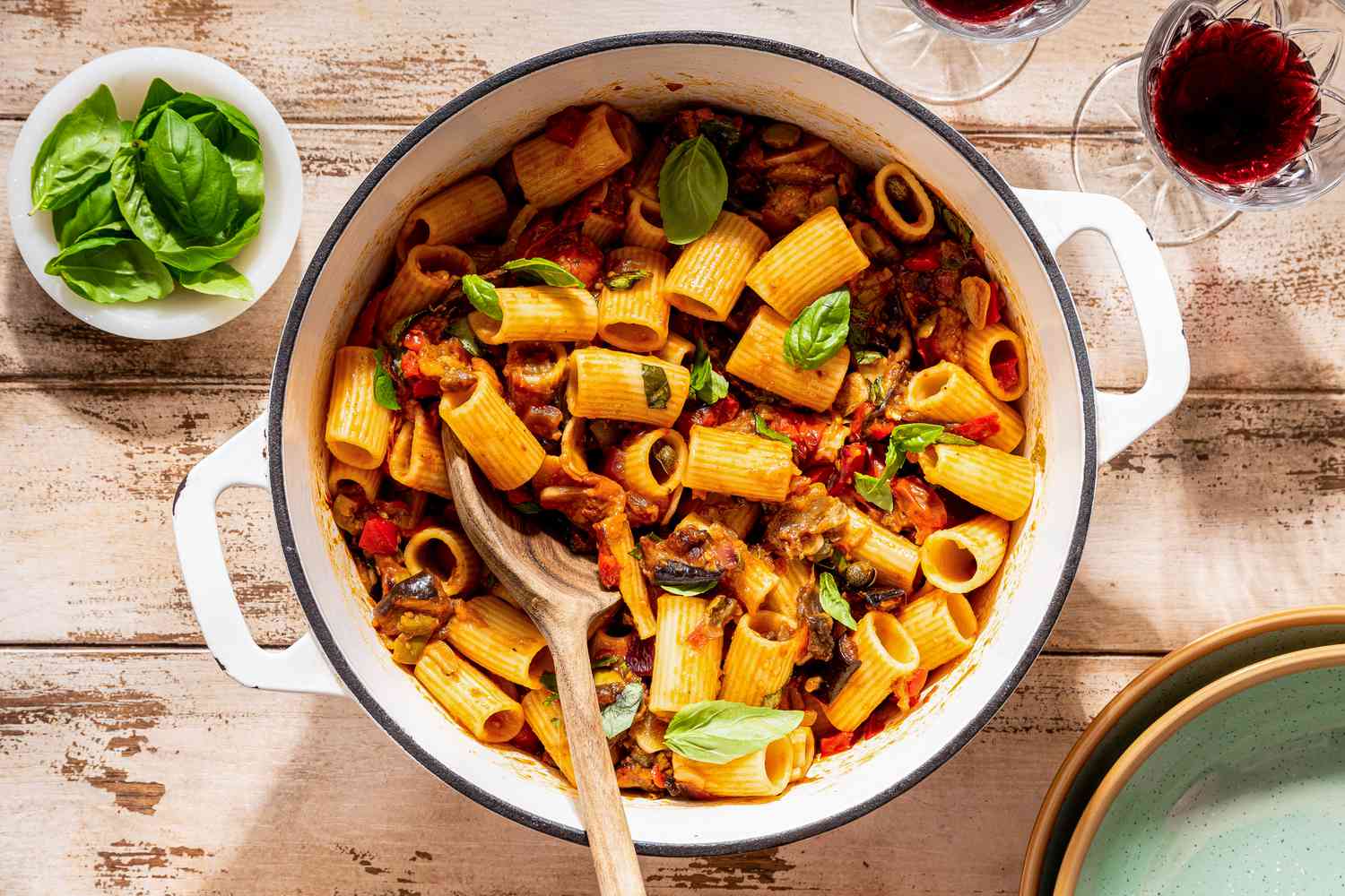 Sicilian caponata pasta in a dutch oven next to a stack of plates, a glass of wine, and a small bowl of basil, all at a rustic table setting 