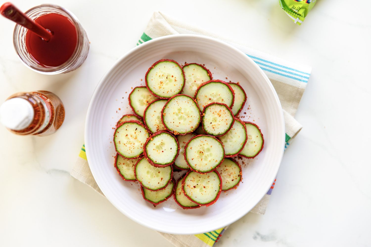 A bowl of cucumber slices seasoned with Tajn placed on a table with a jar of sauce and a spice container nearby