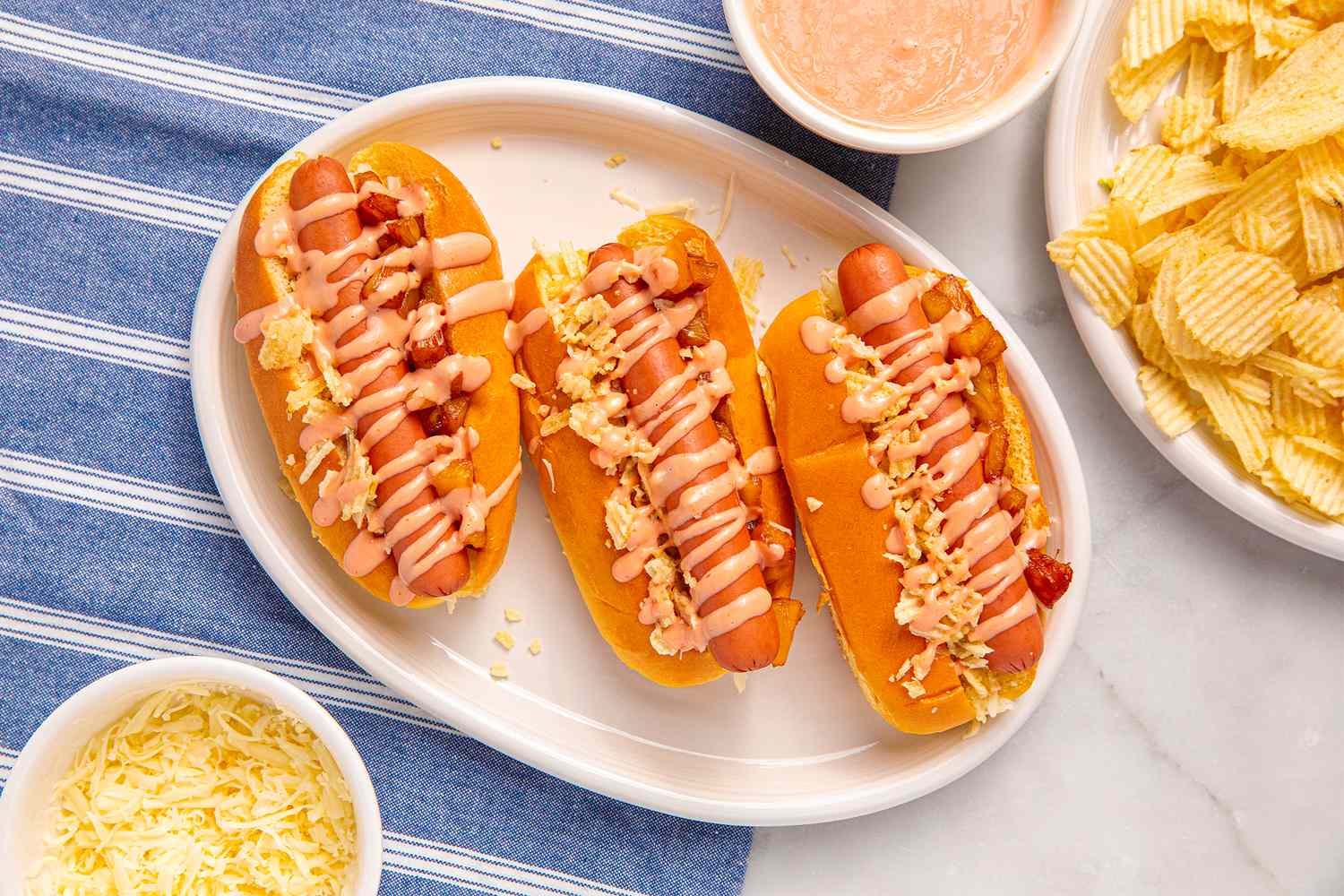 Three hot dogs on a plate with toppings and sauces accompanied by a side of chips and shredded cheese