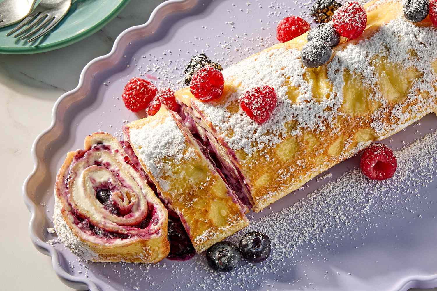 Rolled crepe cake with berry filling dusted with powdered sugar and garnished with fresh raspberries and blueberries on a platter
