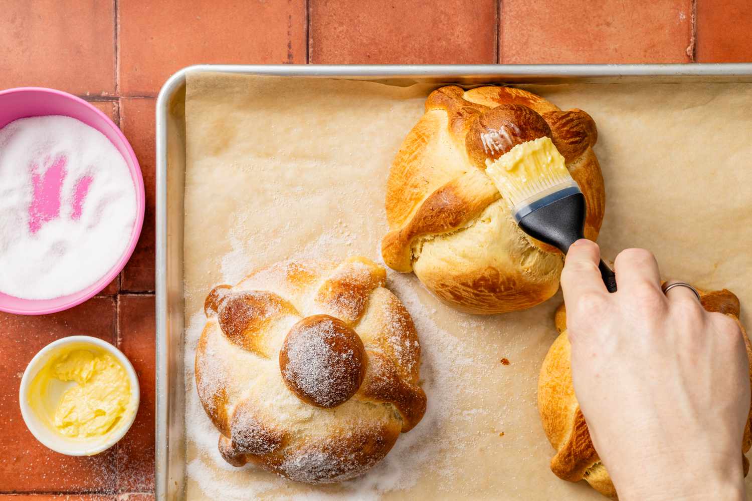 One pan de muerto brushed with butter next to another already topped with sugar (all on a parchment paper lined baking sheet)