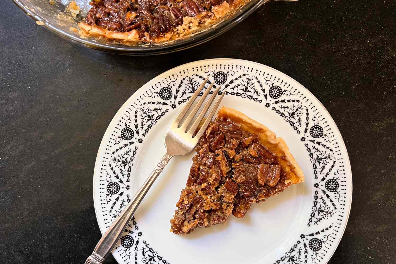 A slice of pecan pie served on a white decorative plate with a fork placed next to it the rest of the pie visible in the background