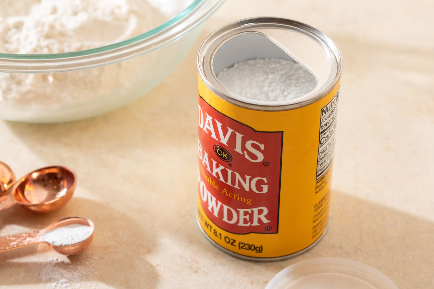Can of baking powder surrounded by tablespoons with some baking powder in one of the spoons and a bowl of flour
