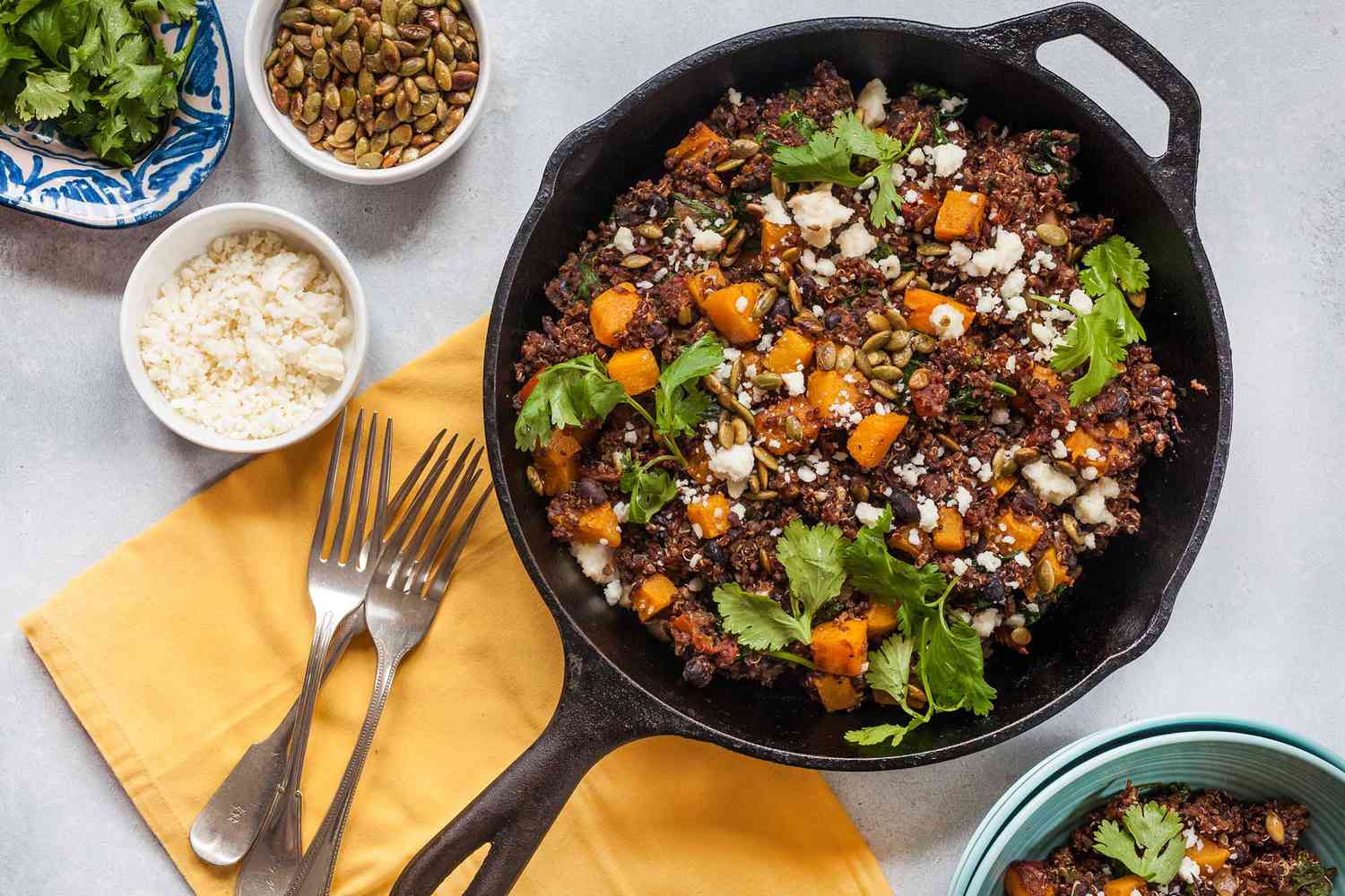 Horizontal view of a large cast iron skillet filled with a butternut squash skillet dinner. Cilantro, quinoa, butternut squash and cotija cheese are visible in the pan. To the left is a yellow napkin with three forks on it. Above the forks are three small bowls. One small bowl of rice, a ramiken of roasted pumpkin seeds and a small platter of cilantro.