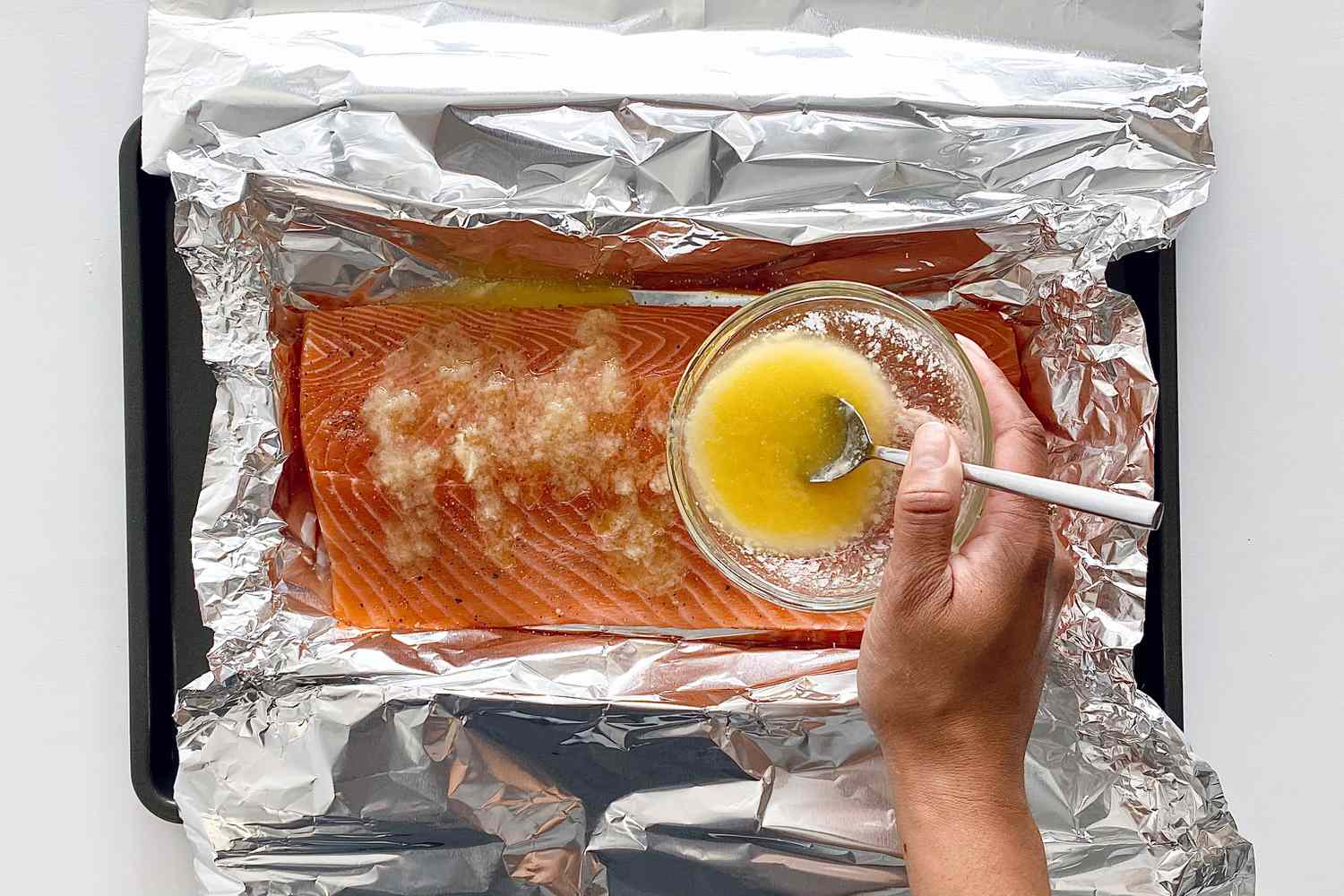Melted butter being poured on an unbaked salmon on a baking sheet