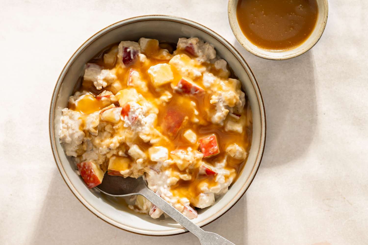 A bowl of caramel apple overnight oats with a spoon next to a small bowl of caramel sauce