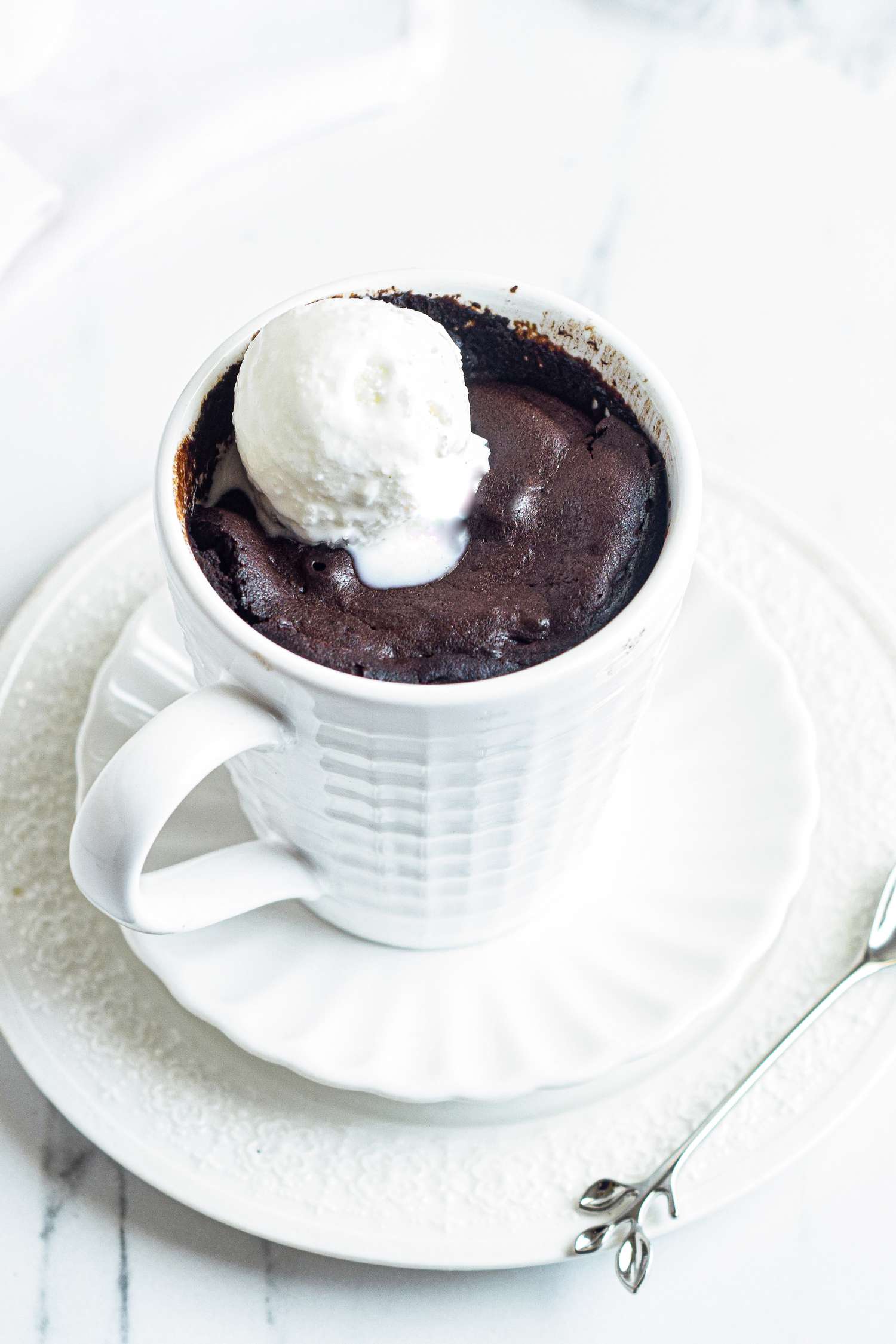 Side view of a brownie in a white mug and topped with ice cream.
