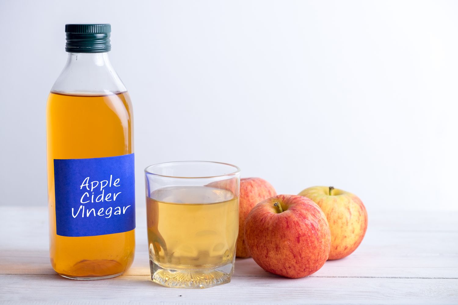 A bottle labeled apple cider vinegar a glass of liquid and three apples on a wooden surface
