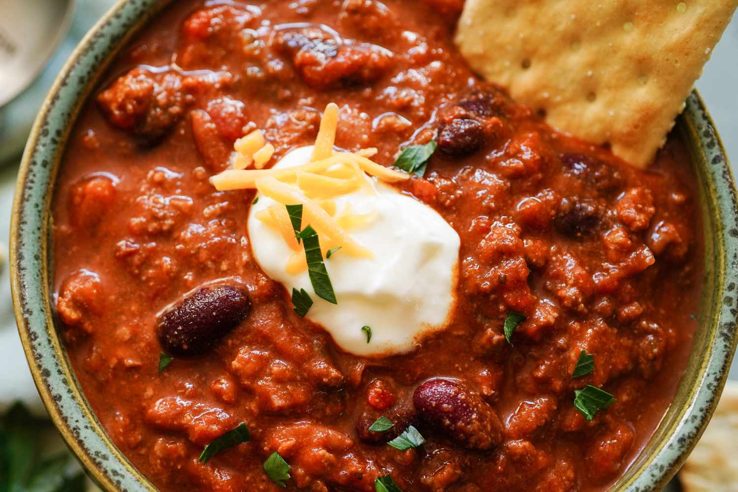 Bowl of chili with kidney beans sour cream shredded cheese and herbs served with a cracker on the side