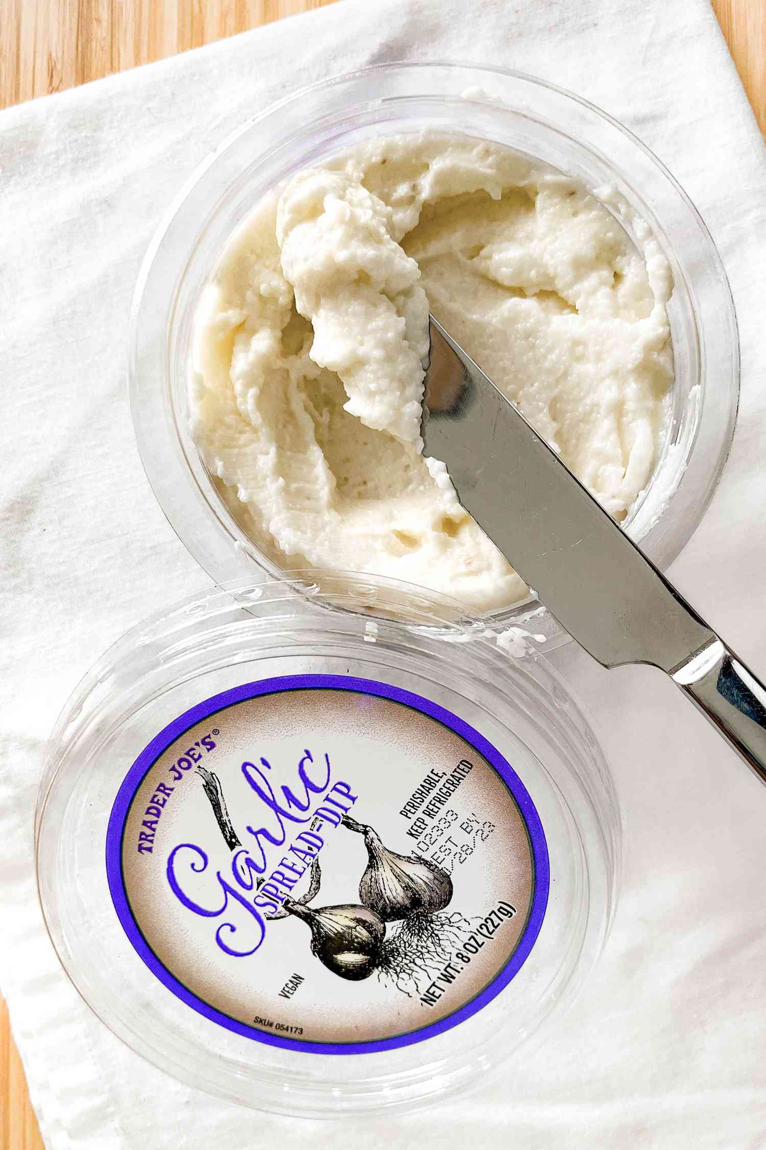 Open tub of Trader Joe's Garlic Spread Dip with a butter knife in the tub (butter knife has some of the dip on it), all on a white kitchen towel
