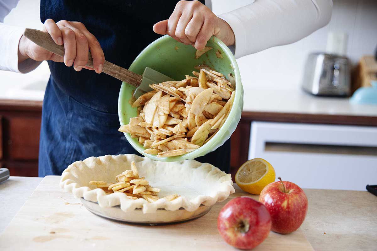 A woman uses a spatula to spoon sliced apples for an easy homemade dutch apple pie into a pie plate. Two apples are on a cutting board nearby.