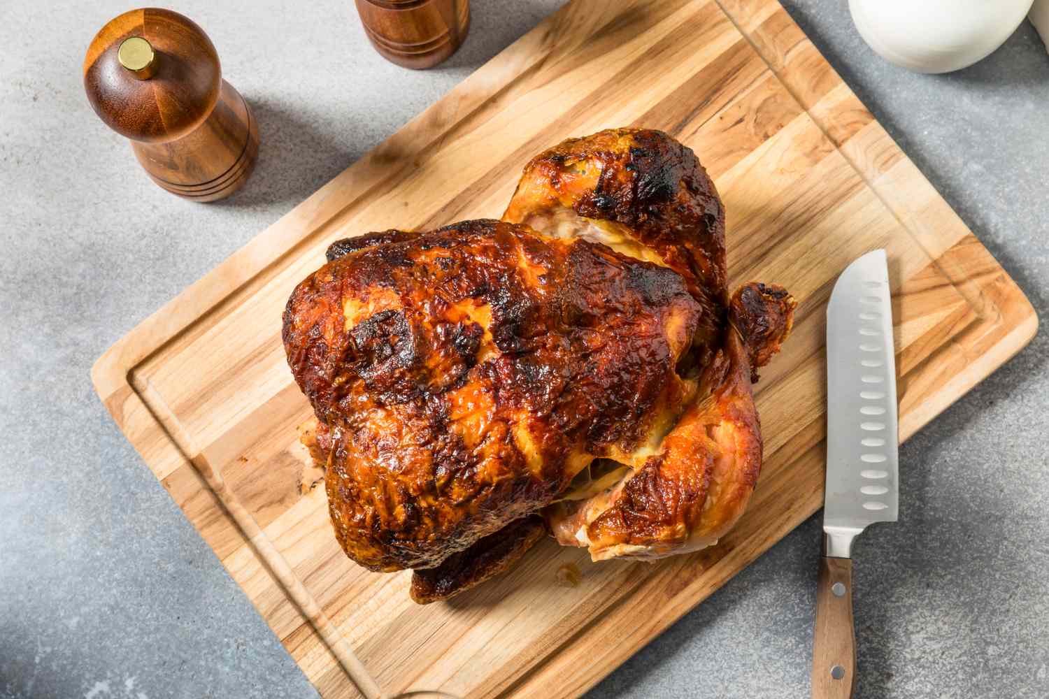 A whole roasted chicken on a wooden cutting board with a knife and condiment containers nearby