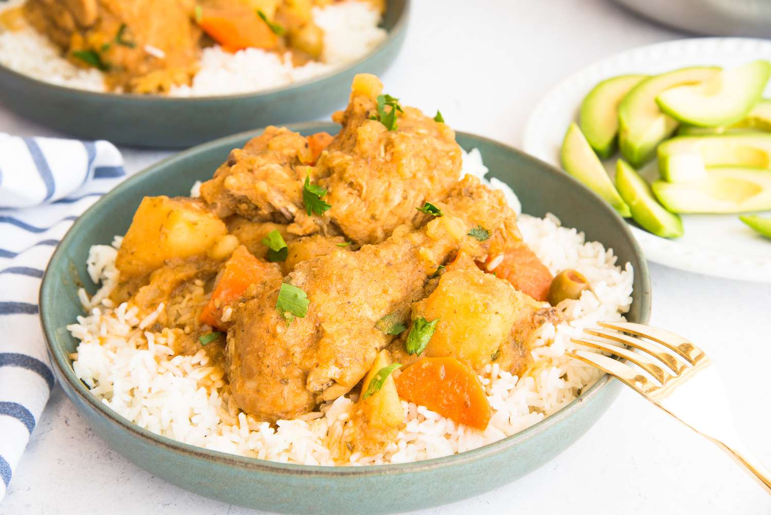 Bowl of Pollo Guisado Served Over Rice Next to a Plate with Sliced Avocado Pieces 
