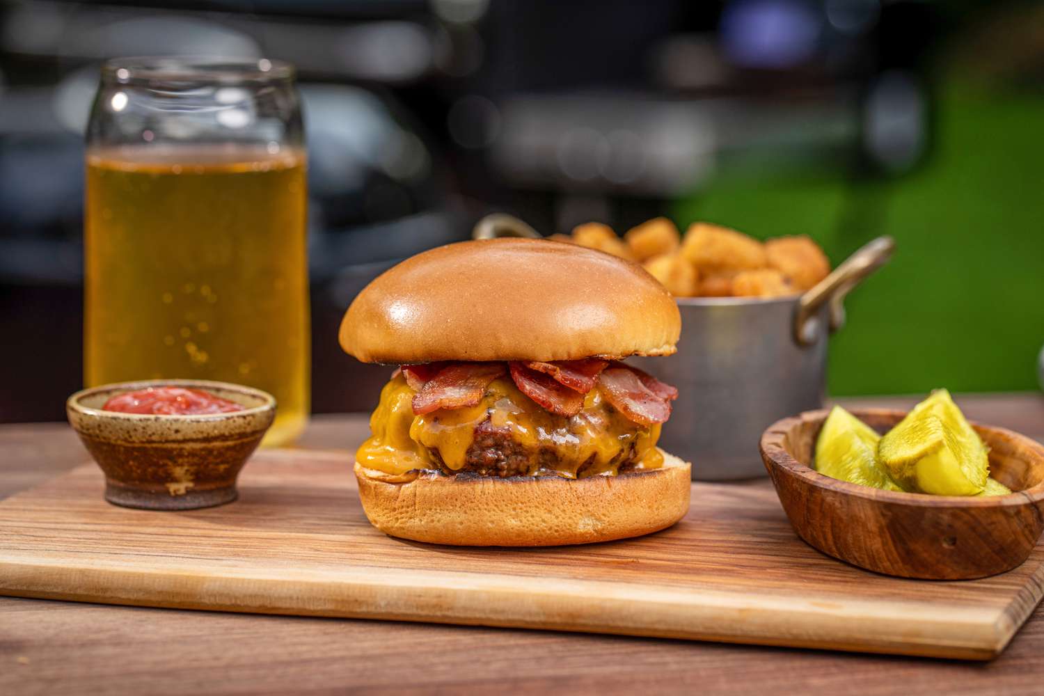 Side view of a perfect bacon cheeseburger outside on a wood cutting board with pickles, beer, and spices set around it.