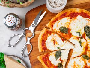 Kitchen shears alongside a partially sliced margherita pizza on a cutting board
