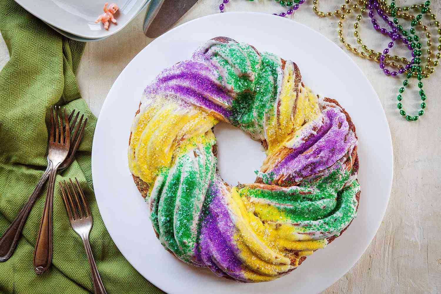 Top view of a homemade king cake on a white plate. Forks and a green linen are to the left. A small plastic baby figurine is above as well as Mardi Gras beads