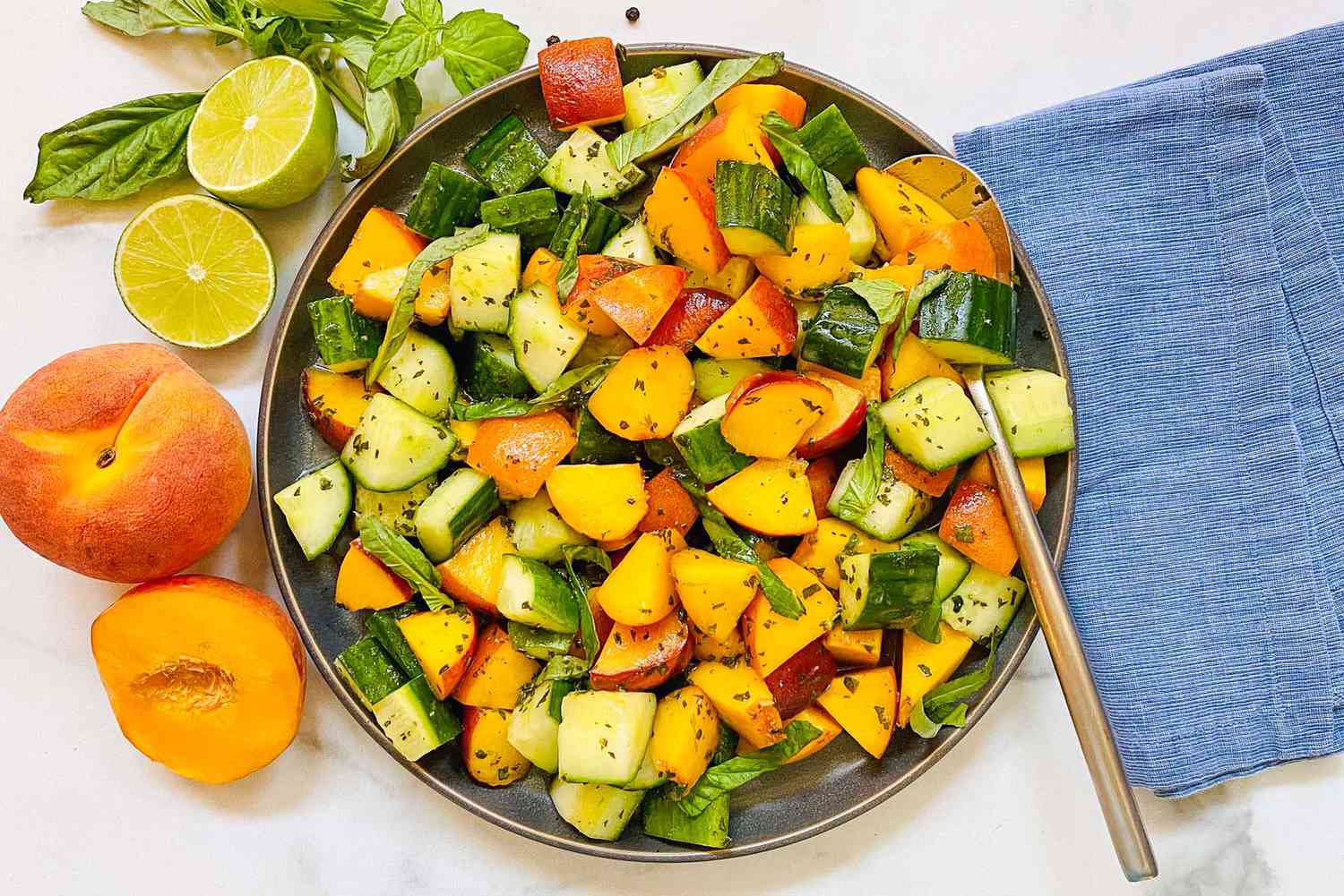 Summer peach and cucumber salad in a bowl with a serving spoon. A blue linen is to the right and peaches, limes and basil are to the left of the bowl.