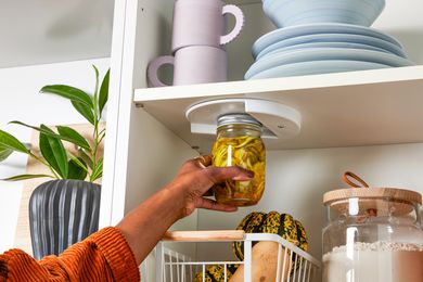Hand Holding a Closed Jar up to a Jar Opener Installed under One of the Cabinet Shelfs. In the Surroundings, a Cabinet Setting With Stacks of Plates, a Salad Bowl, Mugs, a Container of Flour, and a Basket of Fruit and a Potted Plant Next to the Cabinet. 