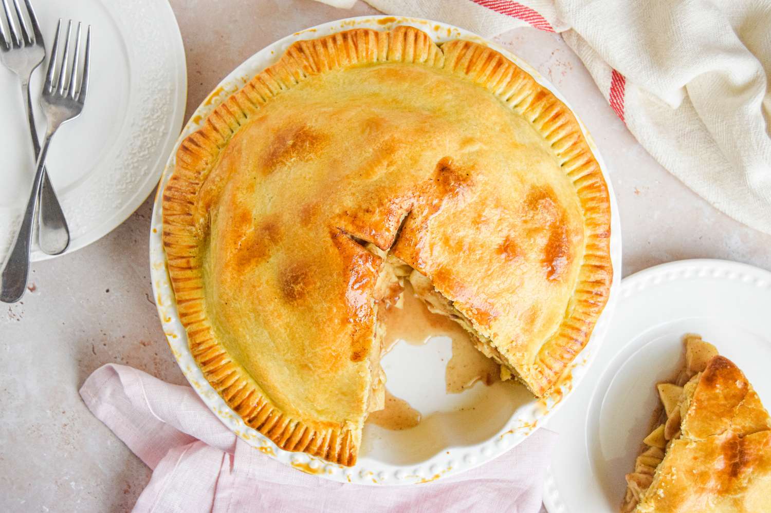 A baked apple pie with a slice removed placed on a plate next to forks and a napkin