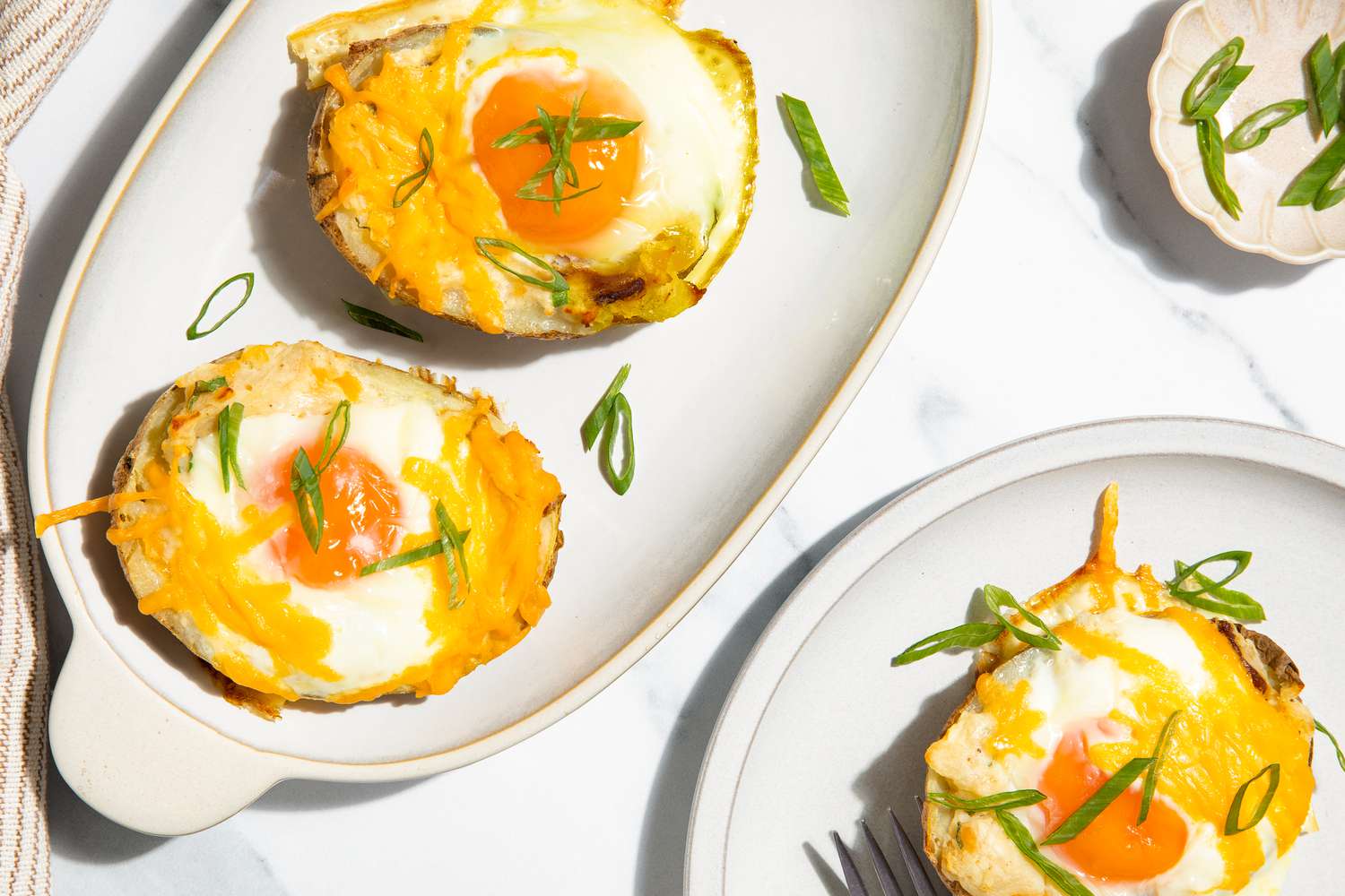 Overhead view of a platter with two loaded potatoes next a plate with one potato, a small bowl of green onions and a cloth napkin all on a marble countertop