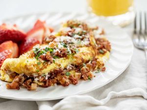 Horizontal image of the best corned beef omelet. The folded omelet is covered in browned corned beef. Grated cheese and parsley are on the top of the omelet. A white cotton napkin is partially under the plate. A fork rests on the napkin which is to the right of the plate. A partial view of a glass of orange juice is above the plate. Halved strawberries are to the left of the omelet on the plate.