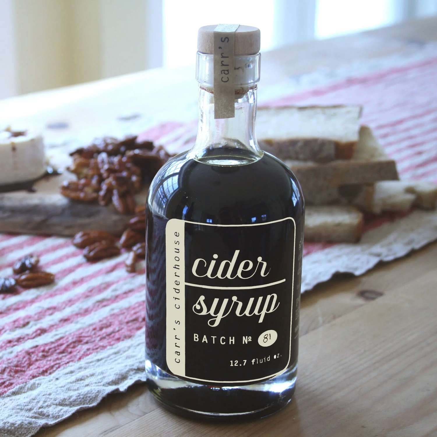 A bottle of cider syrup labeled Carr's Ciderhouse on a wooden table near bread and nuts