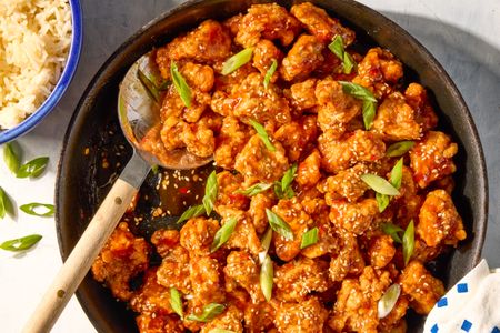 General Tsos chicken dish with sesame seeds and green garnish in a skillet served with rice on the side in a bluerimmed bowl