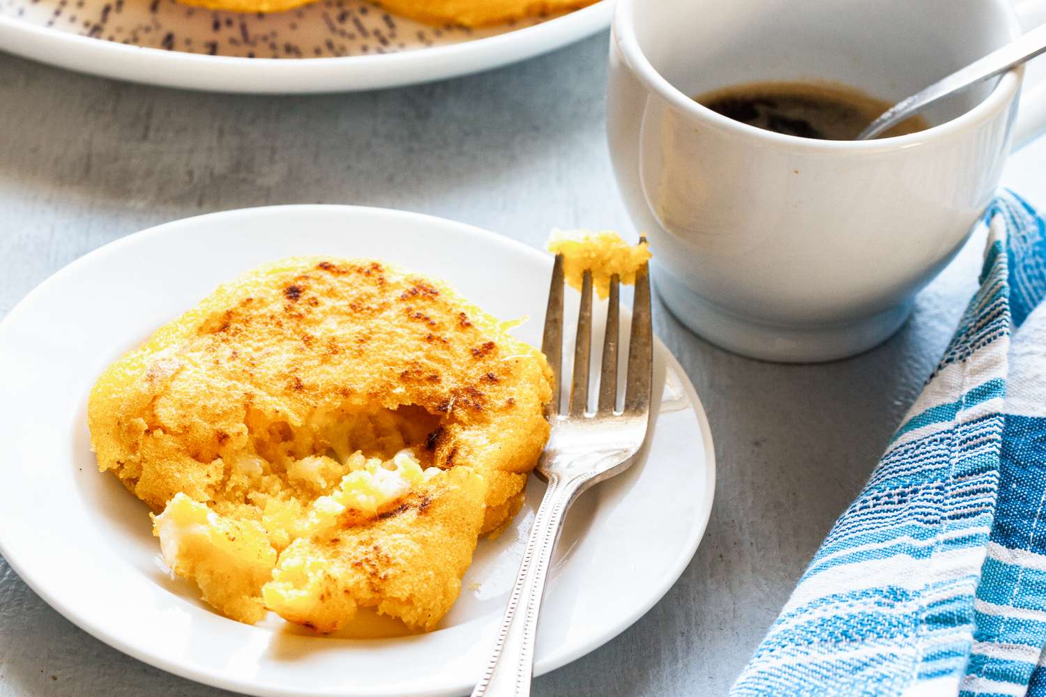 Arepa with cheese on a plate next to a fork and a cup of coffee with a cloth napkin nearby