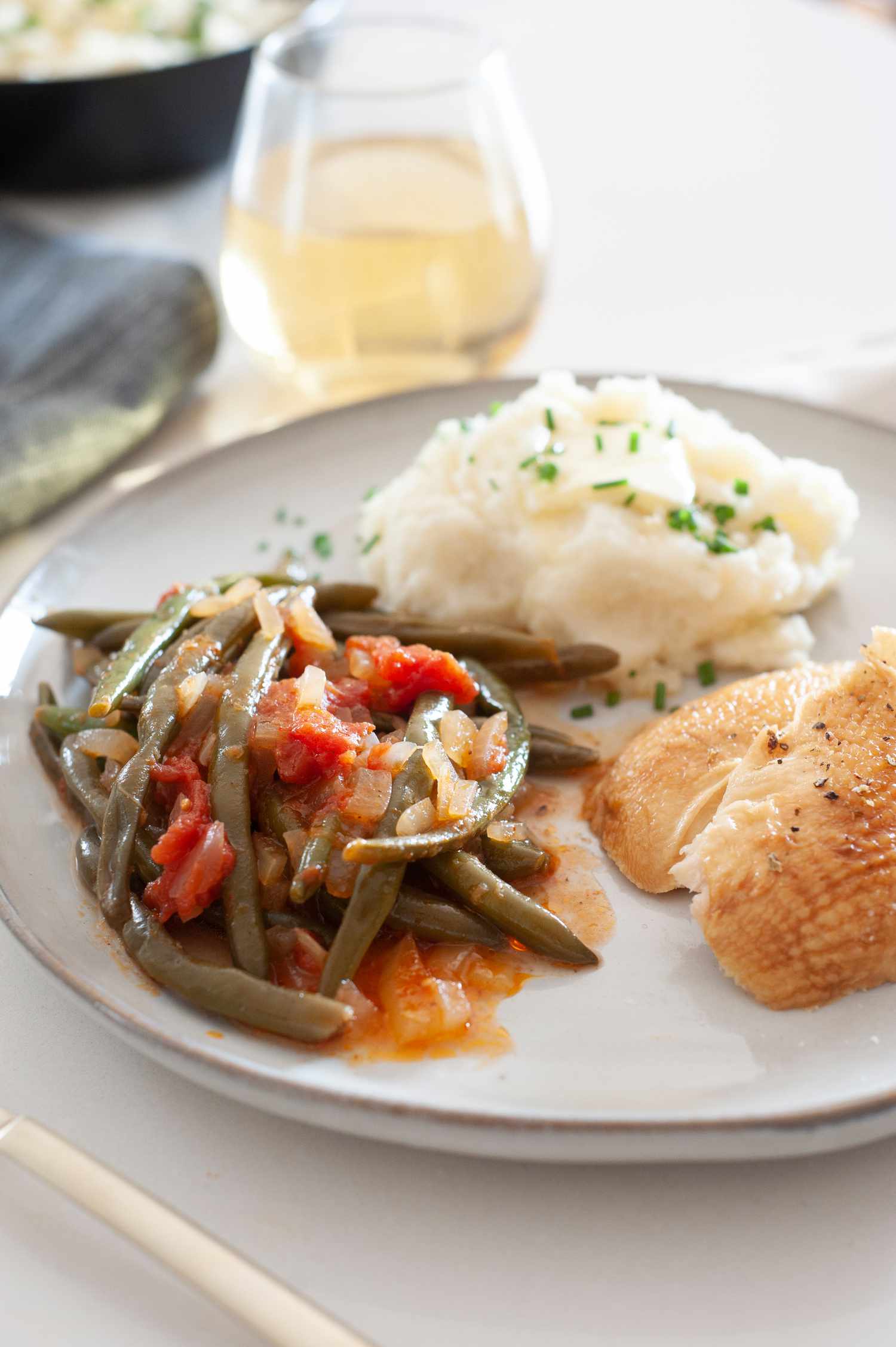 Side view of a plate with green beans in tomato sauce, mashed potatoes and chicken. A glass of wine is behind the plate.