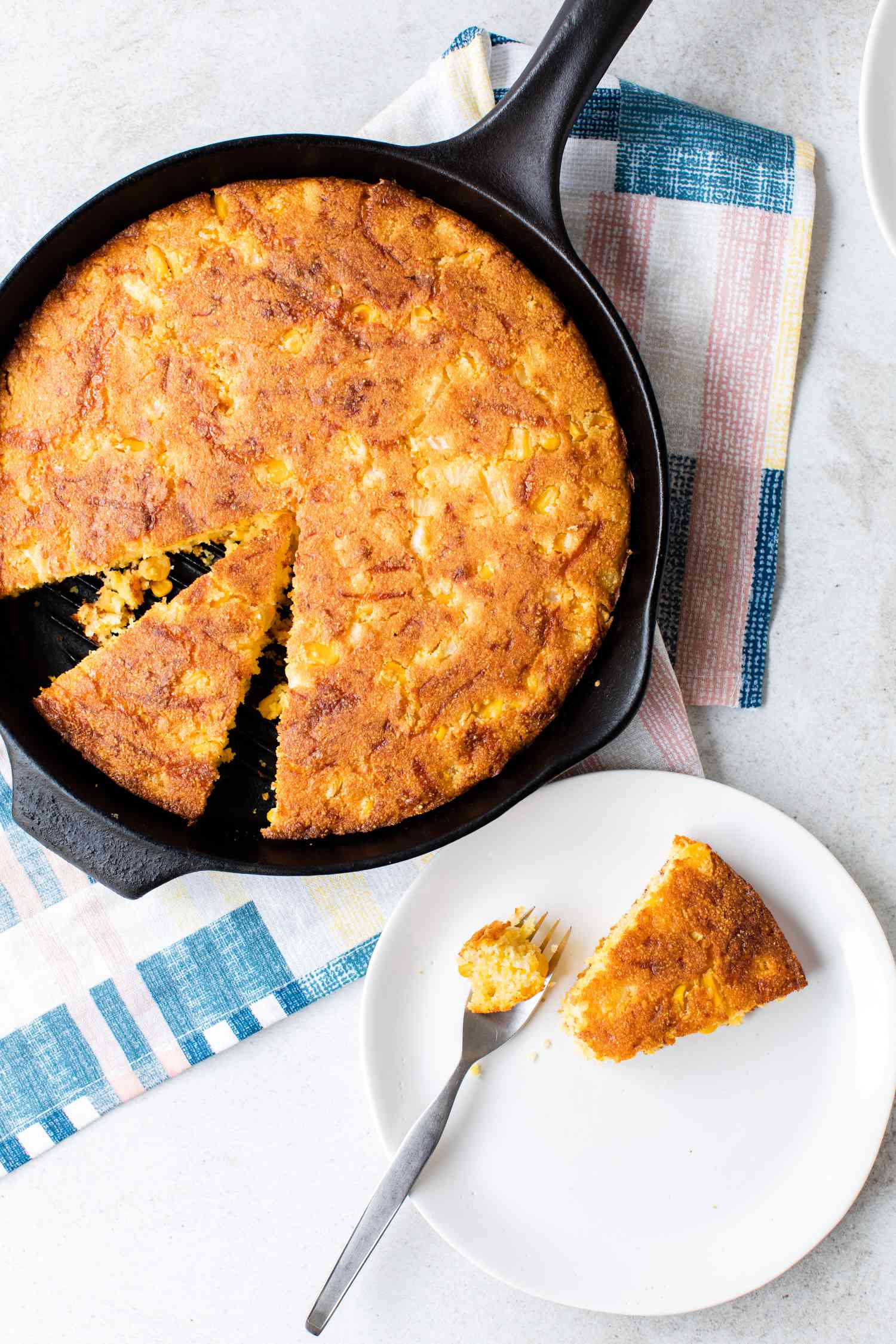A Serving of Cheesy Skillet Cornbread on a Plate with a Fork and the Rest in a Cast Iron Skillet