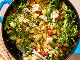 A skillet containing couscous, arugula, roasted vegetables, and cheese crumbles, with a serving spoon