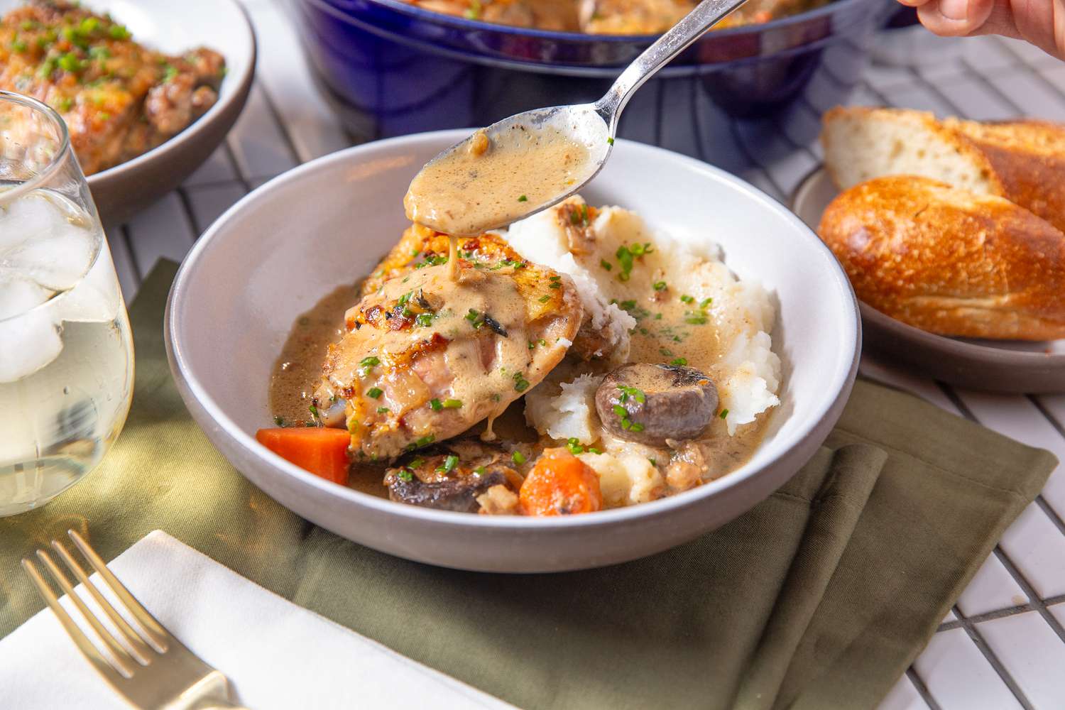 chicken fricassee with mashed potatoes in a bowl with some mashed potatoes and sauce drizzled on it at a table setting with another serving in a bowl, a plate of bread, and more chicken fricassee in a dutch oven