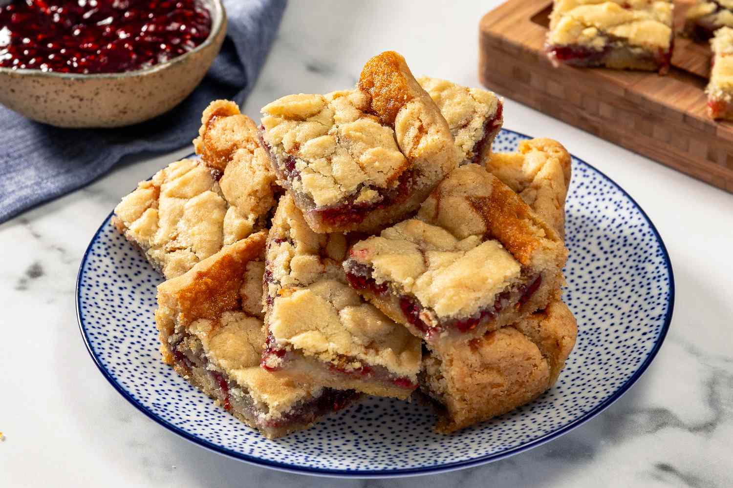 Side view of 2-ingredient jam bar squares piled high on a blue and white plate, with a bowl of jam in the background