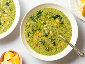 Overhead view of a white bowl of white bean and spinach soup with a spoon next to a plate of bread and bowl of lemon slices