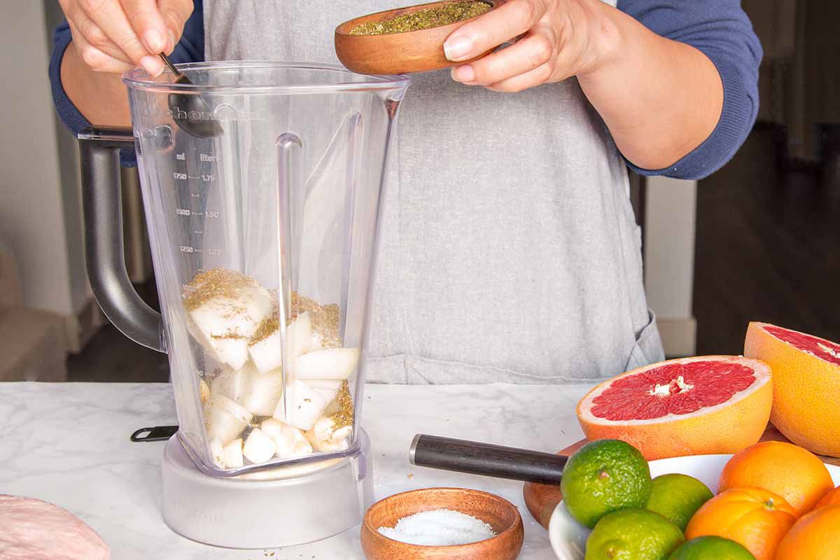 Side view of blender pitcher set on a marble background with limes, oranges and halved grapefruit next to it. A person is putting spices in the pitcher.